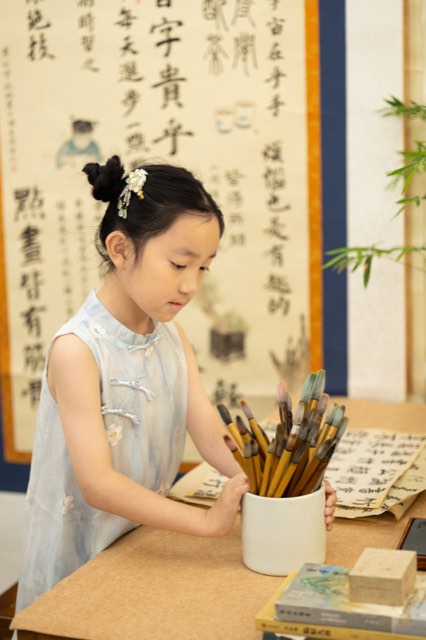 书桌上摆放着毛笔、书帖与书画材料，学员在桌旁整理画笔