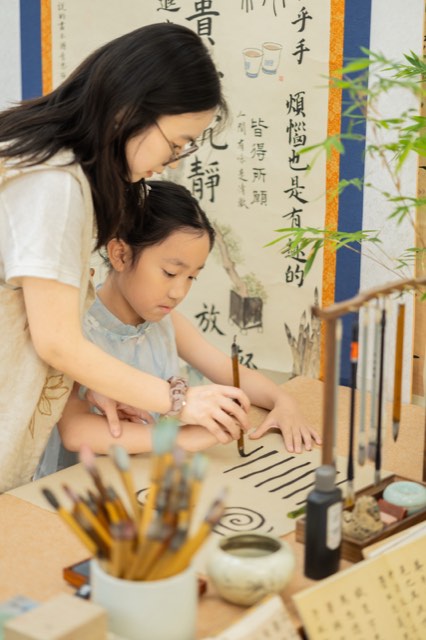 老师在书画教室指导学员用笔，案头有笔架、砚台，墙上悬挂书法