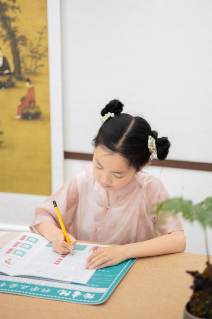身穿粉色上衣的学员坐在木质书桌前，低头专注练习硬笔书写