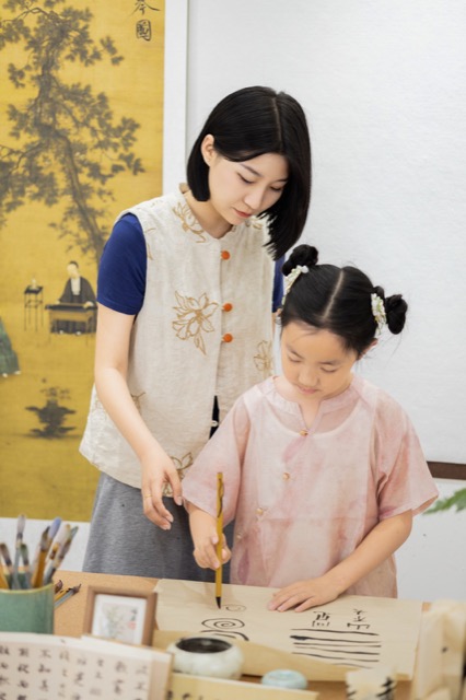 老师在学员身旁指导毛笔握姿与笔画练习，书桌上摆放宣纸和砚台